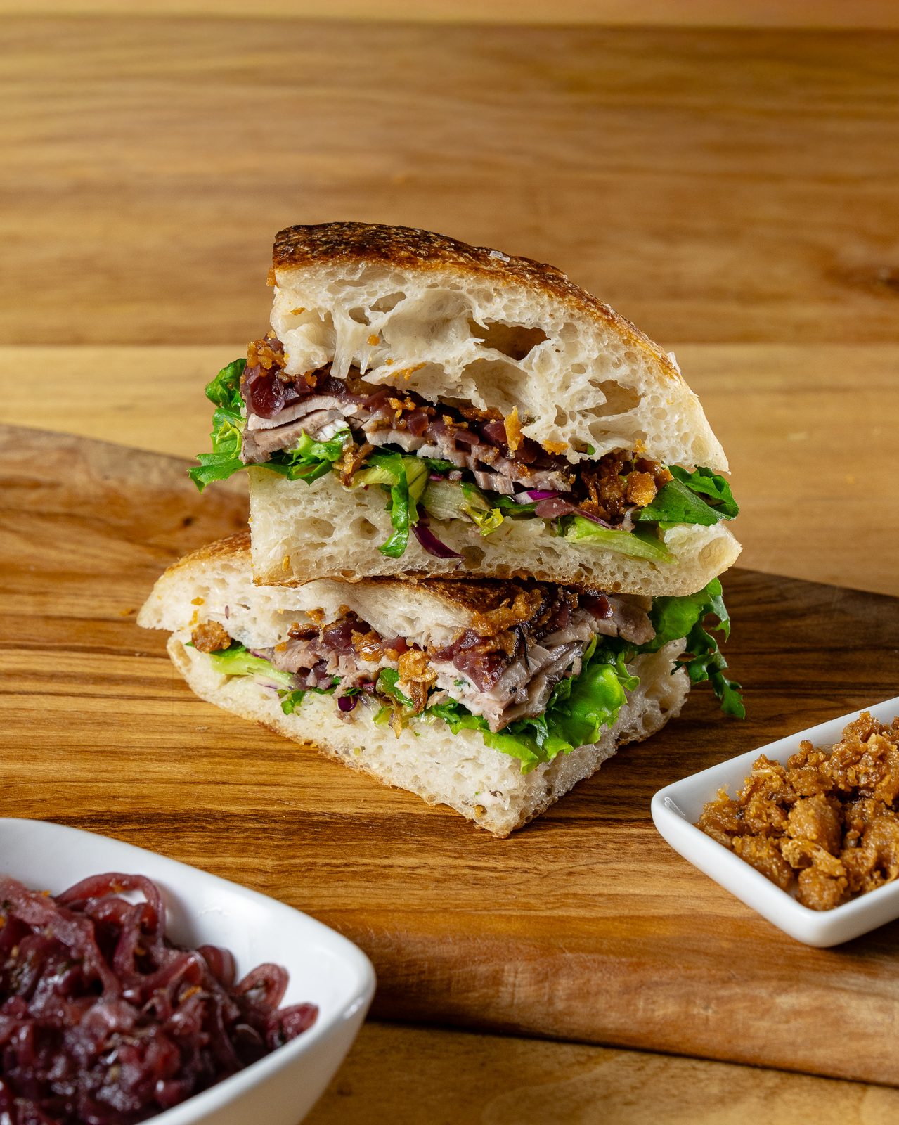 Panino artizanal feliat, cu carne fină, dulceață de ceapă roșie și frunze proaspete, pe tocător de lemn